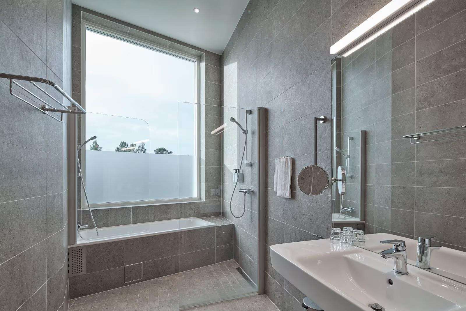 A modern bathroom with a large window, a bathtub, a shower area, and a sink with a mirror.