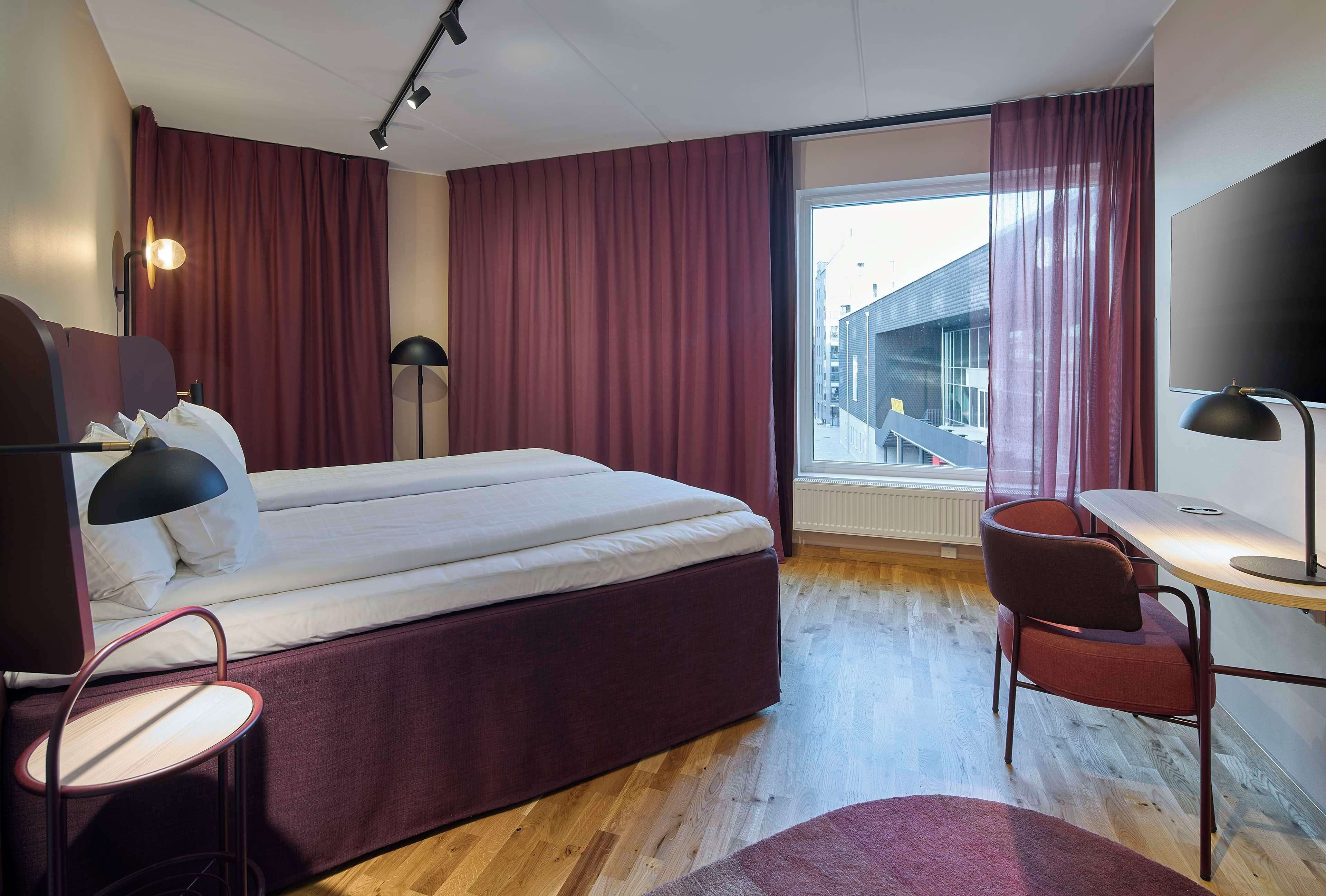 A modern hotel room with a large bed, a desk, and a chair. There is a window with curtains, a radiator, and a lamp.