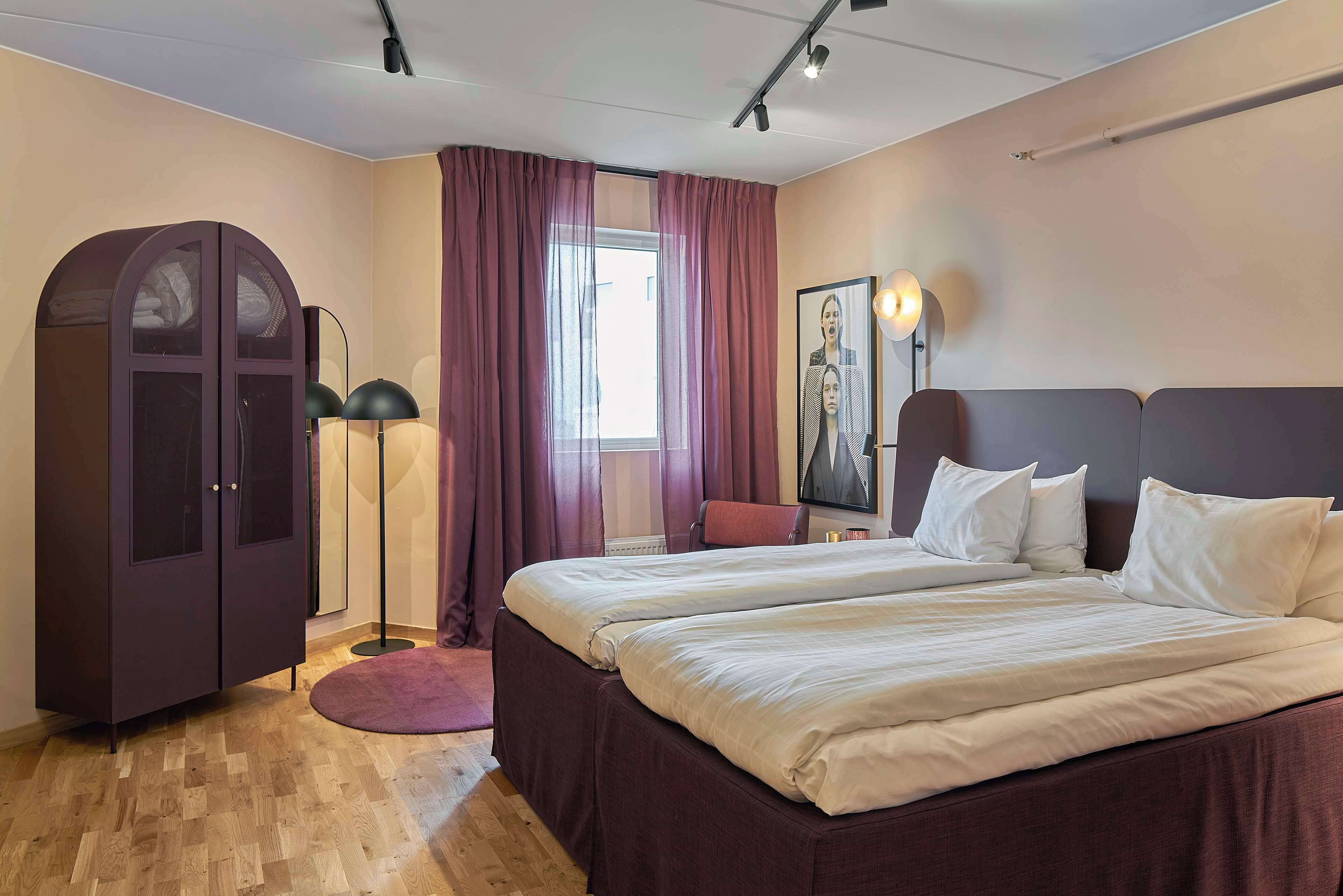 Une chambre d’hôtel avec deux lits, une armoire, une lampe sur pied et une grande fenêtre avec des rideaux.