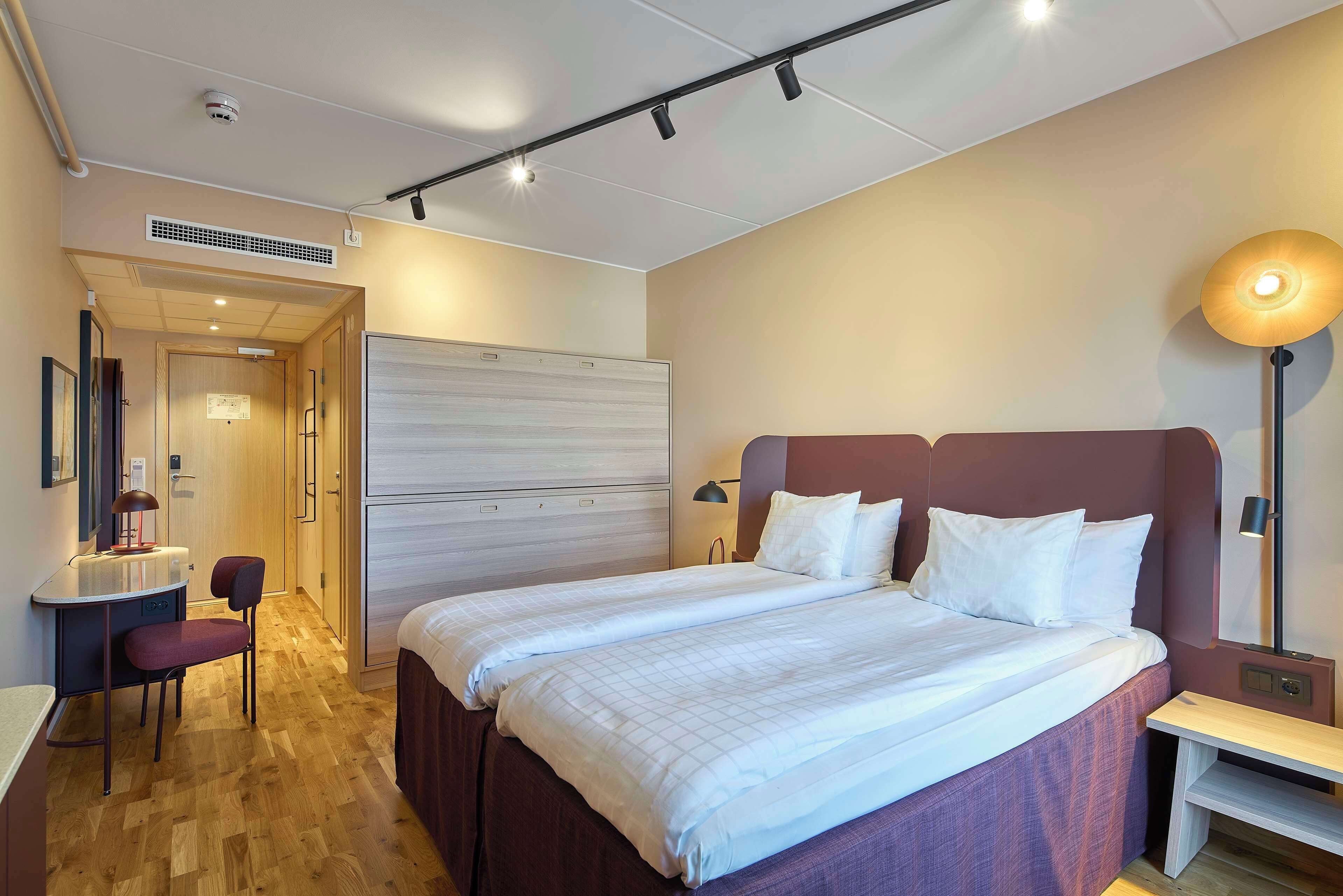 A hotel room with a bed, bedside table, desk with chair, and a lamp.