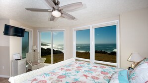 A bedroom with a bed, a ceiling fan, a TV, a chair, and large windows with a view of the ocean. - Promo! Oceanfront Condo with Private Hot Tub, Indoor Pool, Wifi and HDTV! (Depoe Bay)