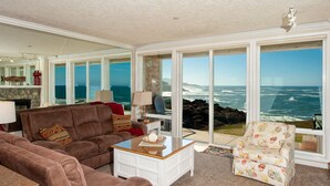A spacious living room with a large window overlooking the ocean, furnished with a brown sofa, a white coffee table, and a patterned armchair. - Promo! Oceanfront Condo with Private Hot Tub, Indoor Pool, Wifi and HDTV! (Depoe Bay)