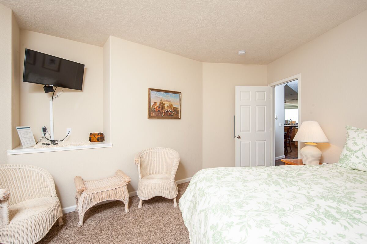A bedroom with a bed, two wicker chairs, a television mounted on the wall, a painting, and a door leading to another room.