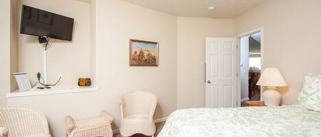 A bedroom with a bed, two wicker chairs, a television mounted on the wall, a painting, and a door leading to another room.