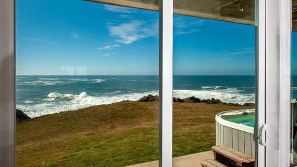 A view of the ocean from a glass door, with a hot tub and wooden bench visible. - Promo! Oceanfront Condo with Private Hot Tub, Indoor Pool, Wifi and HDTV! (Depoe Bay)