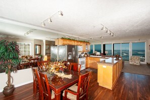 Enjoy meals at the large dining table surrounded by comfortable chairs, with a kitchen and cozy seating area offering a view of the ocean.