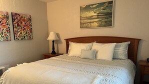 Uma cama bem arrumada com colcha e travesseiros brancos, cabeceira de madeira e mesa de cabeceira com abajur. Há duas pinturas na parede acima da cama.