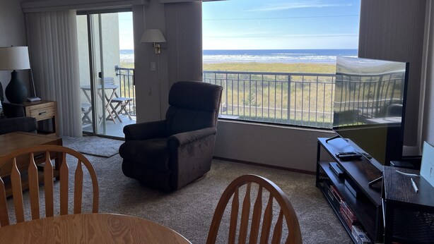 Quarto com uma janela grande que oferece vista para a praia, uma cadeira, uma mesa e um suporte para televisão.