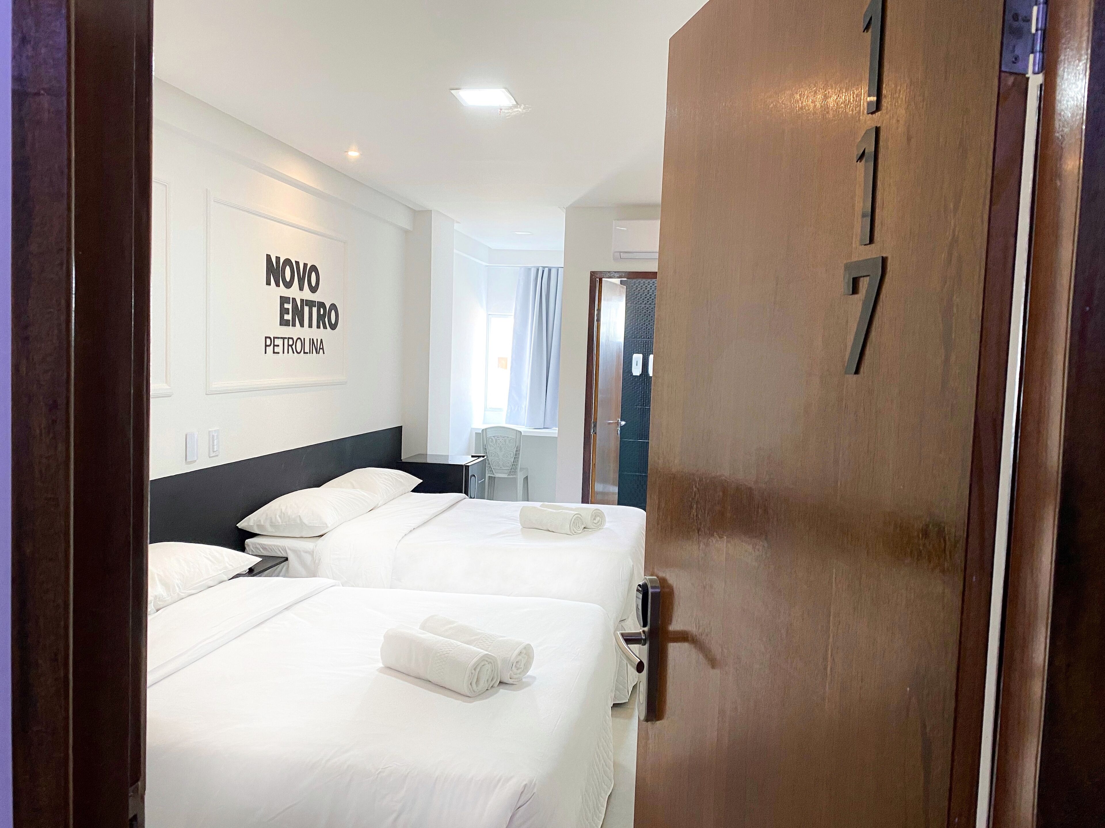 Quarto de hotel com duas camas, uma escrivaninha e uma cadeira. Há um aviso na parede que diz "NOVO ENTRO PETROLINA".