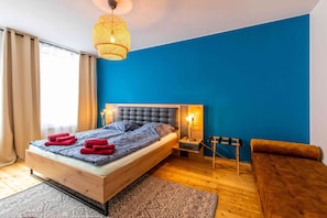 Quarto com cama de madeira, parede azul decorativa, luminária pendente e pequena mesa de cabeceira com abajur.