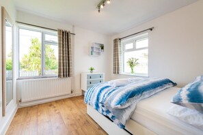 A bedroom with a bed, a radiator, a window with curtains, a bedside table with a plant, and a view of greenery outside.