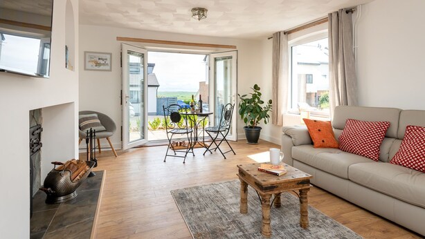 Een ruime woonkamer met een bank, een salontafel en een eethoek met stoelen. Er is een open haard, een groot raam met gordijnen en uitzicht op de omgeving.
