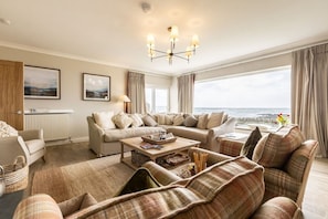 A living room with a sofa set, a coffee table, and a view of the sea. - White Cottage: A large six bed beachside holiday home (Rhosneigr)