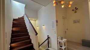 Una escalera de madera con barandilla, una pared blanca con un cartel de 'No Fumar', una pequeña mesa blanca con una botella y una lámpara fijada a la pared.