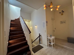 Un escalier avec des marches en bois et une rampe, un mur blanc avec un panneau « Il est interdit de fumer », une petite table blanche avec une bouteille et un luminaire fixé au mur.