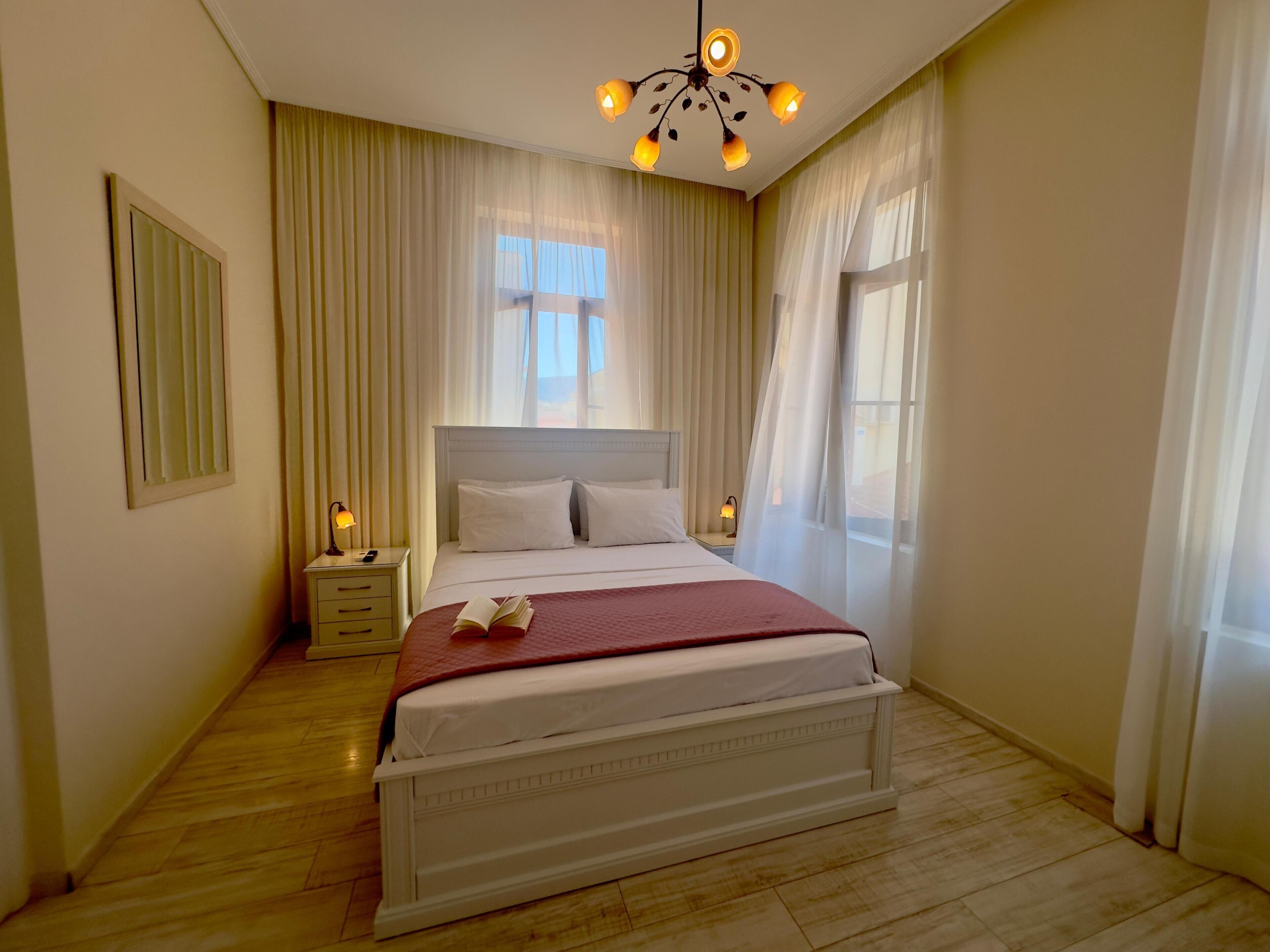 A hotel room with a large bed, a bedside table, a nightstand, a ceiling light fixture, and a window with curtains.