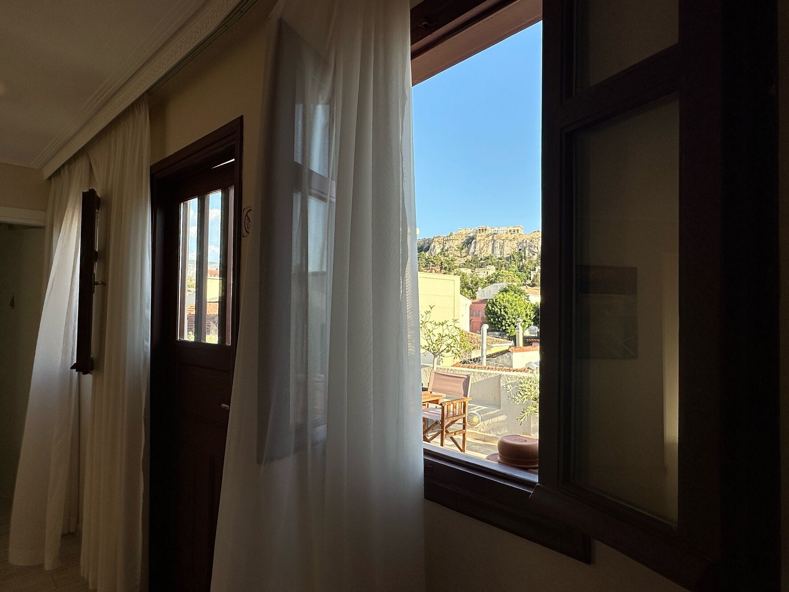 A room with a window overlooking a courtyard with a table and chairs, and a view of a hill with buildings.