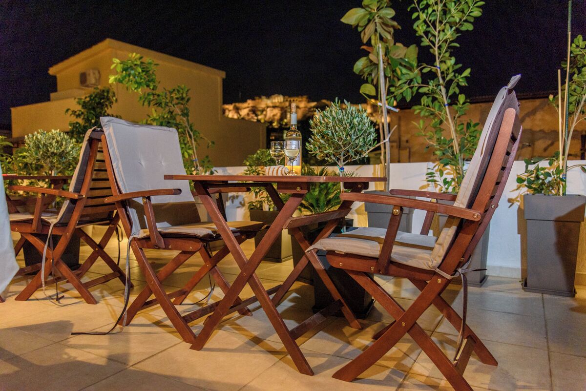 A patio with wooden chairs and tables, a small table with a bottle and glasses, and a building in the background.
