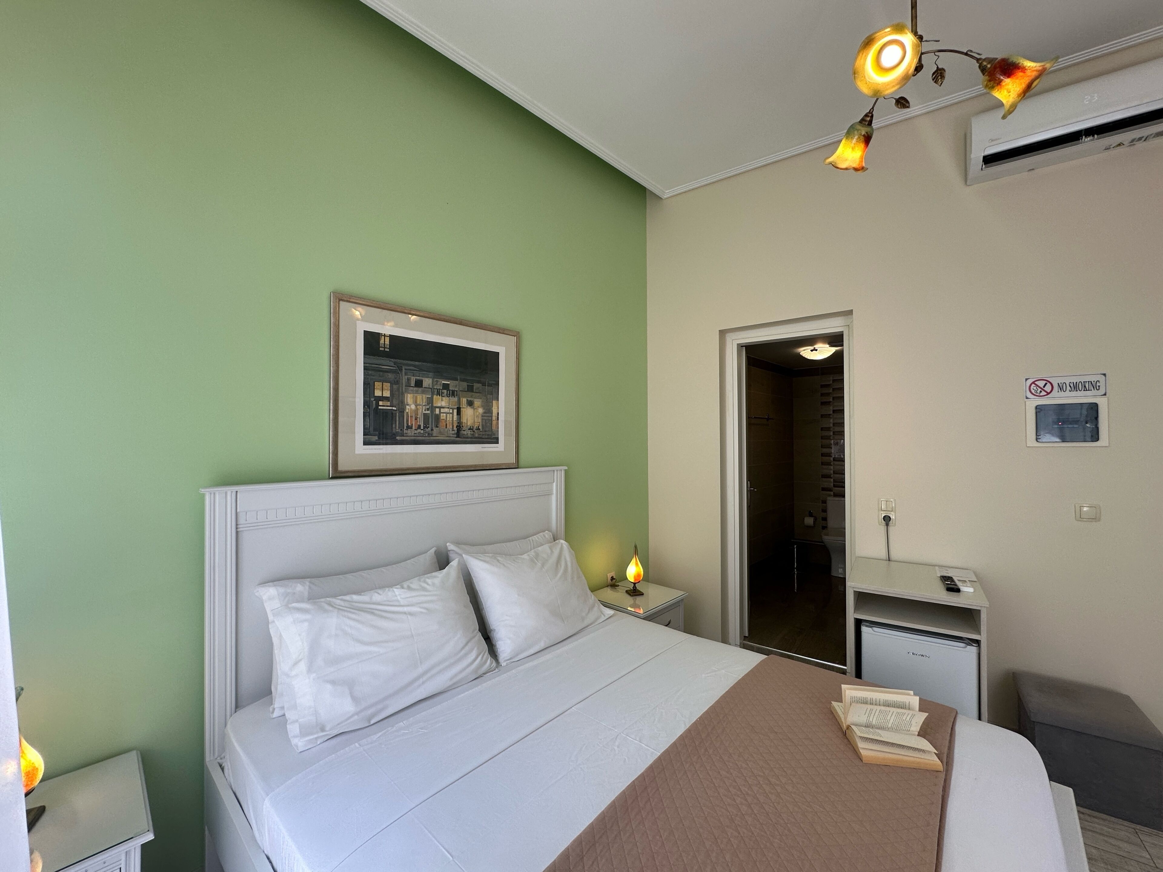 A hotel room with a bed, bedside tables, a framed picture, a shower, a no smoking sign, and an air conditioning unit.