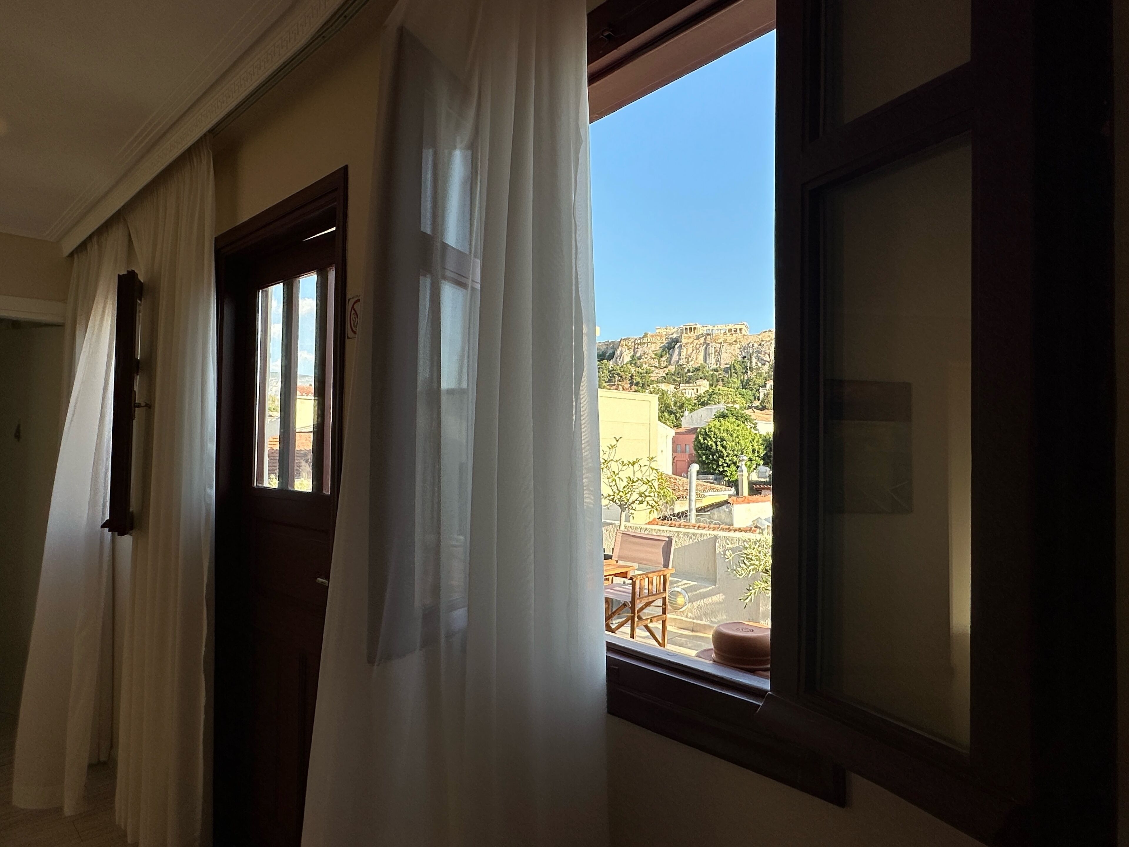 A room with a view of a hill and buildings, a window with curtains, and a door.