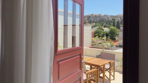Un balcón con mesa y sillas, una puerta roja y vistas a edificios y árboles.