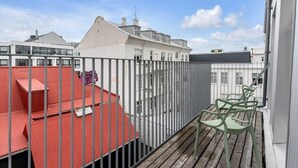 En balkon med gelænder, en bygning med rødt tag, og i baggrunden et hus med hvide vægge og mange vinduer.