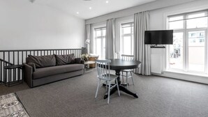 En moderne stue med en grå sofa, et rundt spisebord med stole, et fladskærms-tv og store vinduer med gardiner.