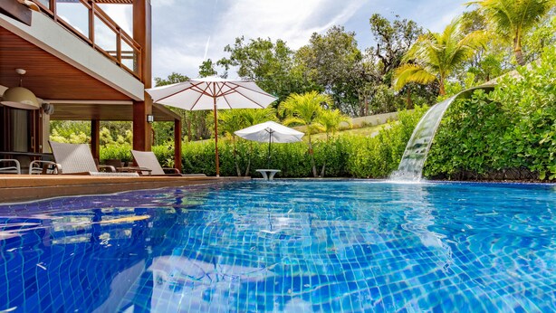Une piscine agrémentée d’une cascade, entourée d’une végétation luxuriante et dotée d’un espace extérieur avec des sièges et des parasols.