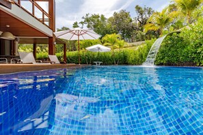 Une piscine agrémentée d’une cascade, entourée d’une végétation luxuriante et dotée d’un espace extérieur avec des sièges et des parasols.
