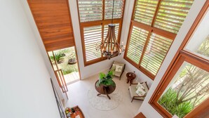 Une pièce aux hauts plafonds, dotée de grandes fenêtres, d’un lustre central, d’une table ronde agrémentée d’une plante, et d’un coin salon avec des coussins.