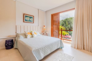 A bedroom with a large bed, a small table, a chair, and a window with curtains. - PF13 Casa 05 Suítes - Praia do Forte (Mata de São João)