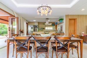 A modern dining area with a chandelier, wooden chairs, and a kitchen in the background. - PF13 Casa 05 Suítes - Praia do Forte (Mata de São João)