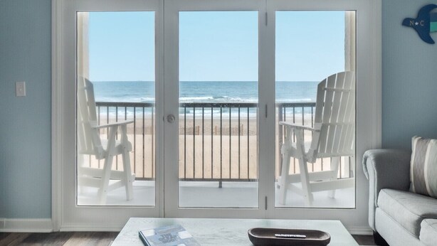 A room with a view of the beach, featuring a sliding glass door, two white chairs, a white sofa, and a coffee table with a magazine and a remote control.