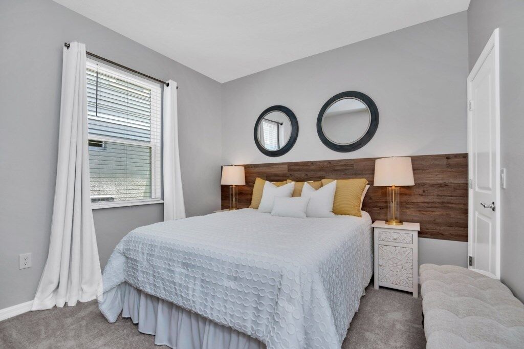 A bedroom with a bed, two bedside lamps, a nightstand, a mirror, and a window with curtains.