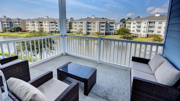 A balcony with wicker furniture and a view of multi-story buildings across a body of water. - 4827 Magnolia Lake Drive, #302 3 Bedroom Condo by RedAwning (Myrtle Beach)