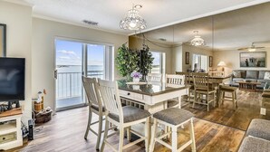 A spacious living area with a dining table, chairs, a TV, and a balcony with a view of the sea. - Harbour Island 403l 2 Bedroom Condo by RedAwning (Ocean City)