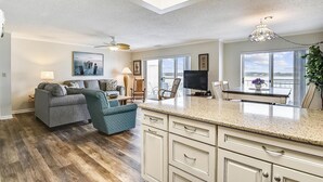 A spacious living area with a grey sofa, teal armchair, and a dining table with chairs. There is a kitchen area with white cabinets and a large window offering a view. - Harbour Island 403l 2 Bedroom Condo by RedAwning (Ocean City)