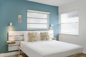 A bedroom with a bed, two bedside tables, a wall-mounted light, a macrame wall hanging, and two windows with blinds. - GROVE27-3 | relax near boho chic neighborhood (Miami)