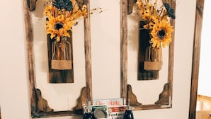 Ein Holztisch mit rustikalem Design, zwei gerahmte Vasen mit Trockenblumen, eine Wasserflasche, eine Tasse, ein Kürbis und eine Karte.