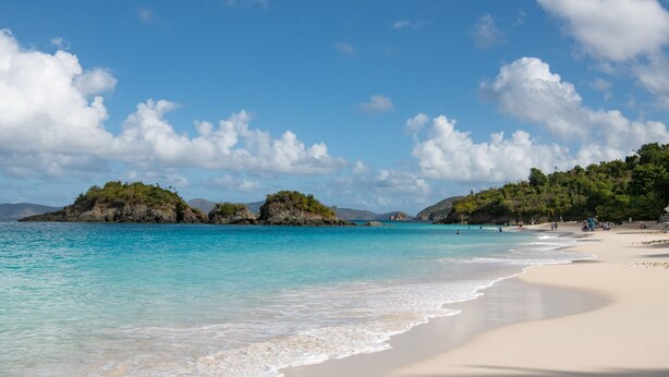 Une plage à l’eau turquoise et limpide, avec un rivage sablonneux et des îlots rocheux couverts de végétation.