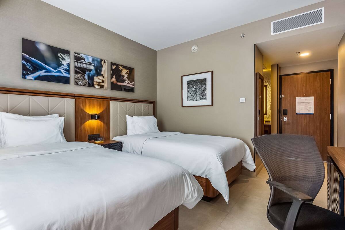 A hotel room with two beds, a desk, and a chair. There are wall-mounted artworks and a framed picture.