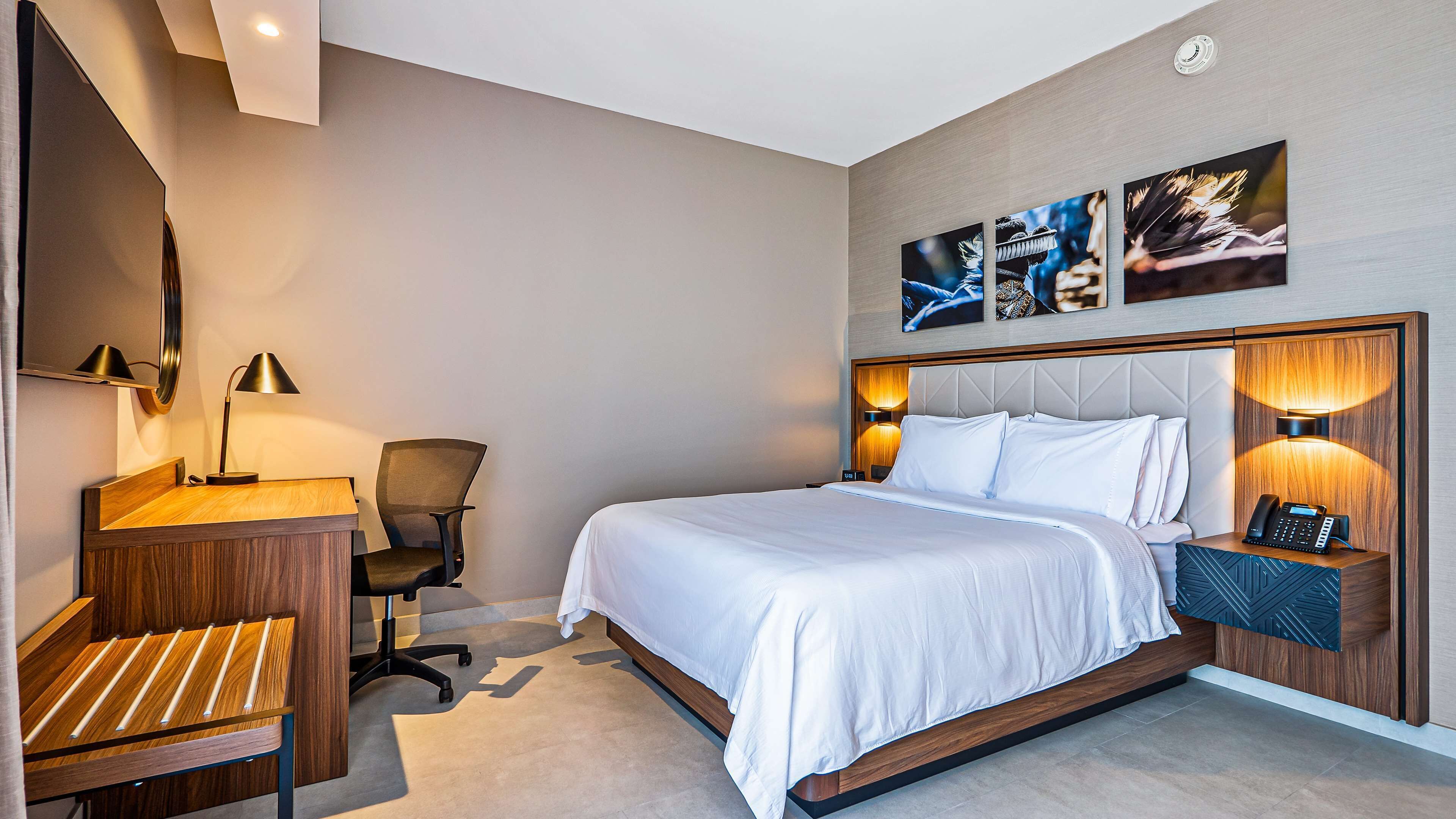 A hotel room with a bed, desk, chair, and artwork on the wall.