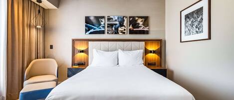 Habitación de hotel con una cama grande, una silla y una mesita de noche con una lámpara. Encima de la cama hay tres cuadros enmarcados en la pared.