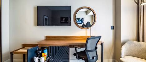 A modern hotel room with a wooden dining table, a black office chair, a sofa, a TV mounted on the wall, and a mirror.