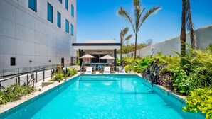 Una piscina con sillas de descanso y sombrillas, rodeada de vegetación exuberante y un edificio moderno.