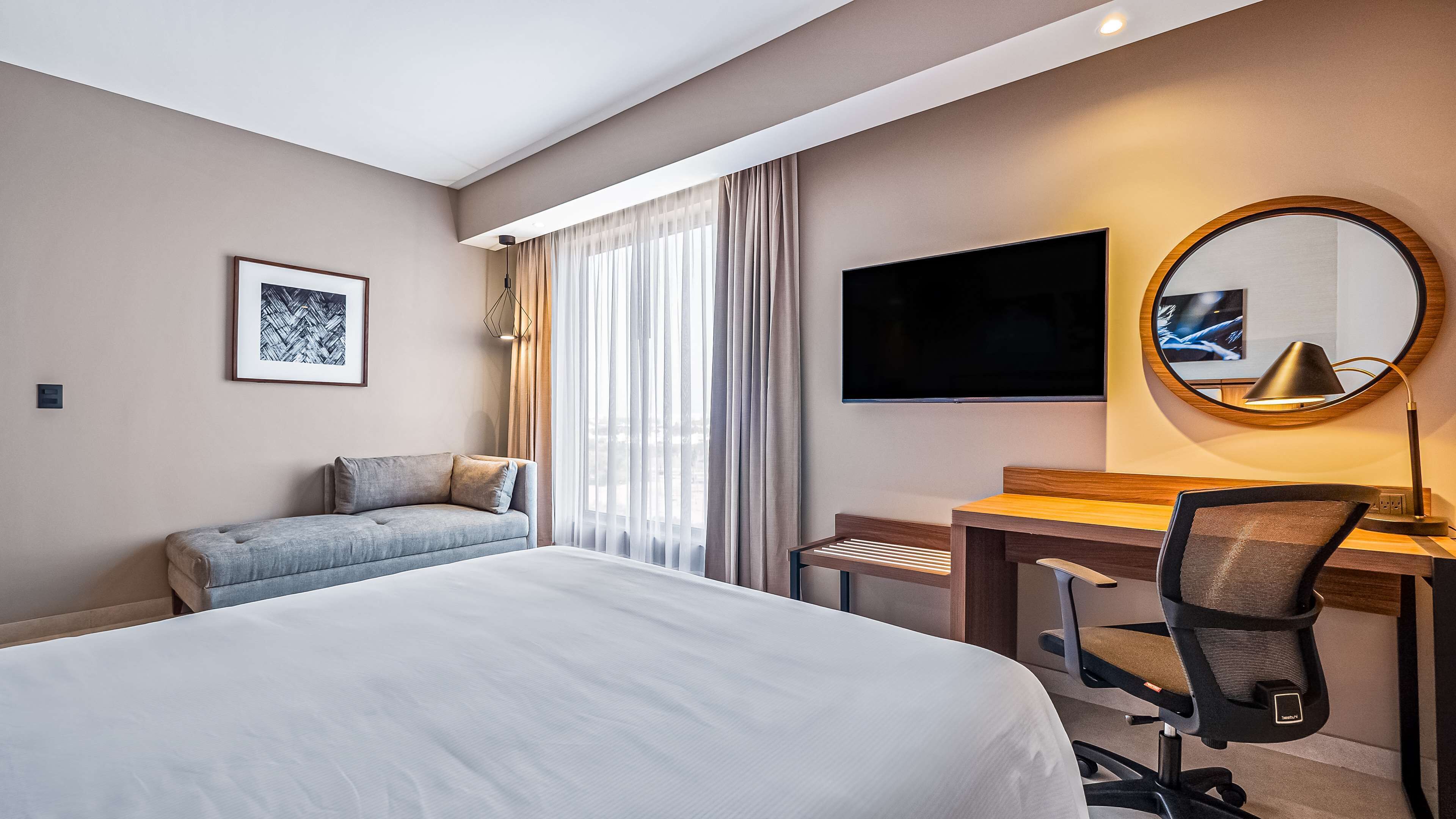 Habitación de hotel con cama, escritorio, silla, televisión, un cuadro enmarcado y ventana con cortinas.