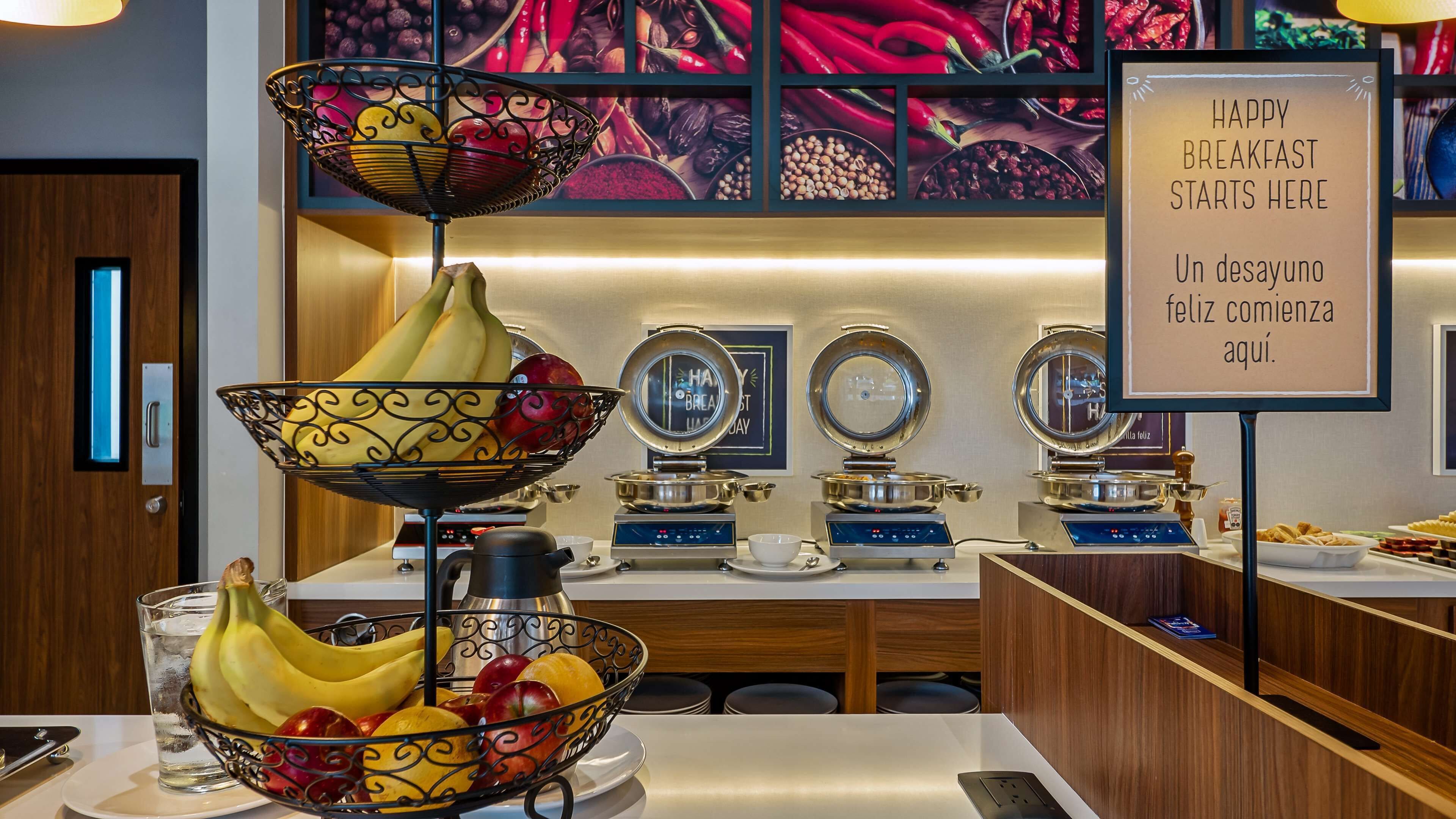 A breakfast buffet with fruit bowls, hot plates, and a sign indicating breakfast starts here.