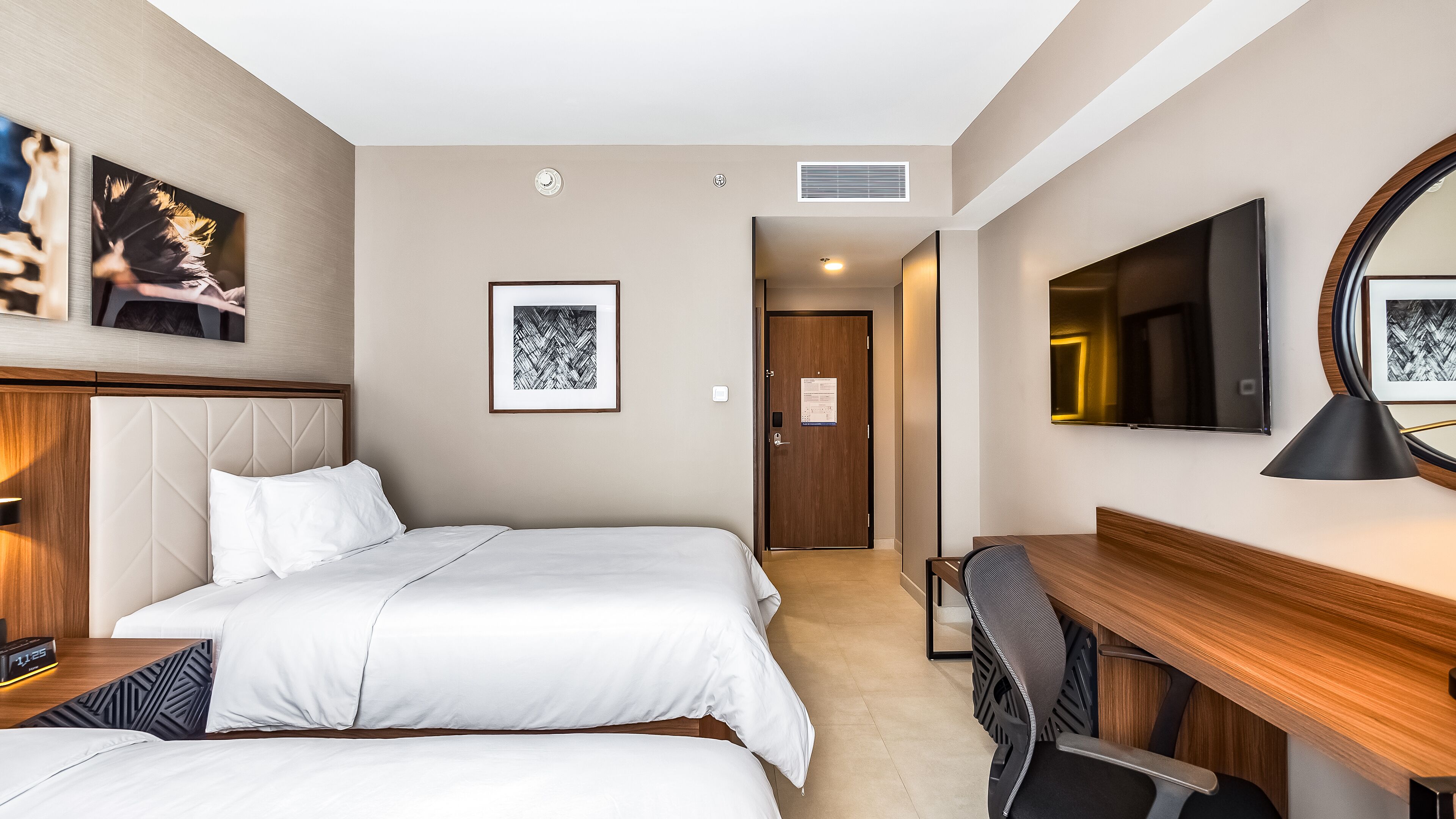 A hotel room with two beds, a desk, a chair, a TV, and artwork on the walls.