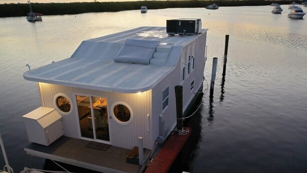 Una casa flotante amarrada en aguas tranquilas, con cubierta techada y un pequeño espacio de estar al aire libre.