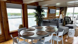 Un salón espacioso con una mesa redonda, sillas blancas, un sofá gris y vistas a una masa de agua a través de grandes ventanales.